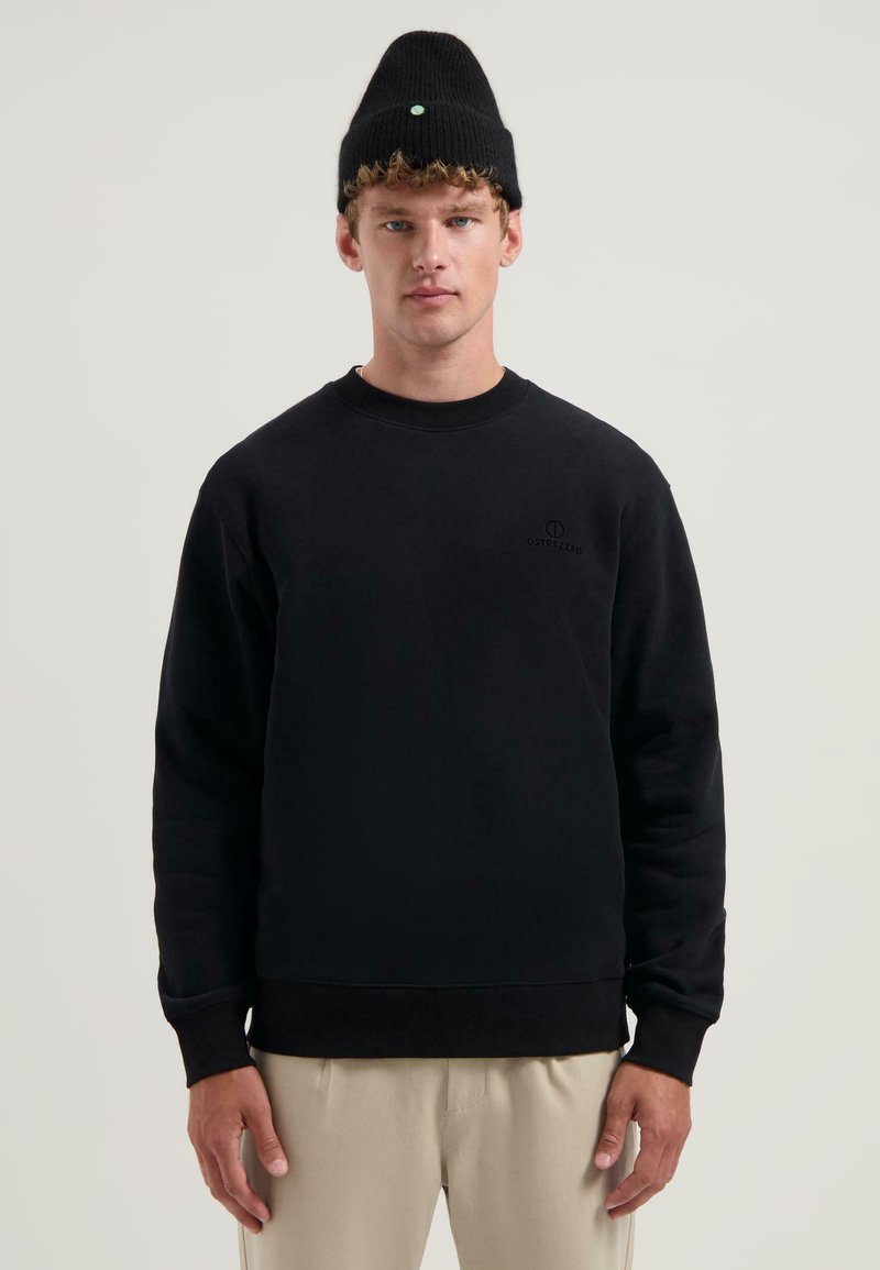 Czarny sweter z okrągłym dekoltem, z dyskretnym logo na piersi. Model nosi czapkę typu beanie z podwiniętym mankietem oraz zielonym akcentem.