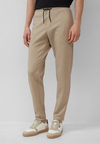 Beige Sporthose mit elastischem Bund und schwarzem Kordelzug. Glatte Textur, schmaler Schnitt, seitliche Nahttaschen, kombiniert mit weißen Sneakern.