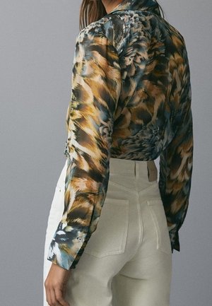 Woman wearing a long-sleeve blouse with an abstract tiger print and high-waisted light beige pants, shown from behind.