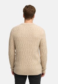 Beige neulepusero, jossa on teksturoitu muotoilu, pitkät hihat, pyöreä pääntie ja ribattuja hihansuita. Viimeistelty suoran helman kanssa.