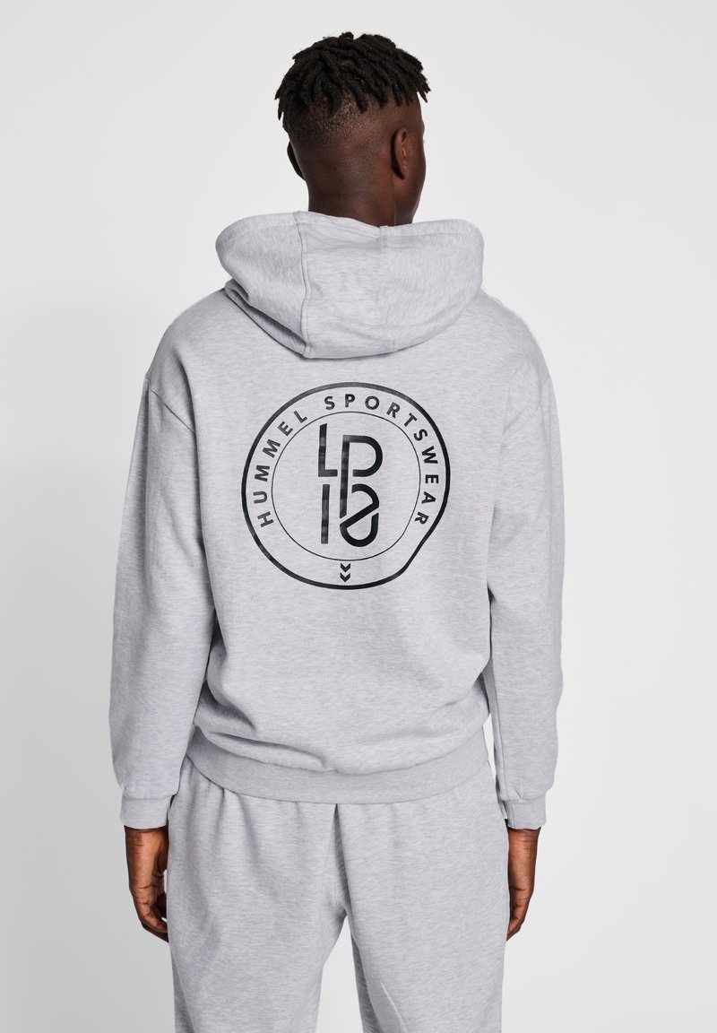 Homme portant un sweat à capuche gris clair et un pantalon de survêtement, dos tourné vers la caméra, avec un logo circulaire noir "Hummel Sportswear" sur le sweat.
