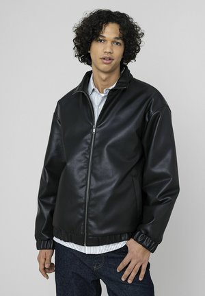 Blouson aviateur en similicuir noir avec fermeture éclair frontale, poignets côtelés, coupe décontractée, superposé à une chemise blanche à rayures bleues.