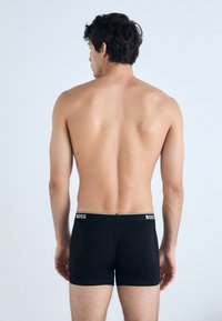 Schwarze Boxershorts mit einem weißen Logo-Bund, figurbetontem Design und weicher Textur, von hinten auf einem neutralen Hintergrund präsentiert.