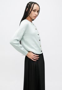 Világoskék kötött kardigán arany gombokkal, kerek nyakkivágással és hosszú ujjakkal, fekete pliszírozott szoknyával párosítva. Simogató tapintású, laza fazon.