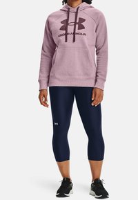 Vaaleanpunainen huppari, jossa on suuri musta Under Armour -logo, yhdistettynä laivastonsinisiin capri-housuihin. Näkyy teksturoitu kangas, narut ja kenguru-tasku.