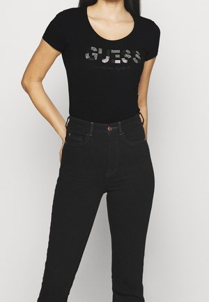 Kvinde iført sort T-shirt med korte ærmer og "GUESS"-logo samt højtaljede sorte jeans, stående med én hånd på hoften.