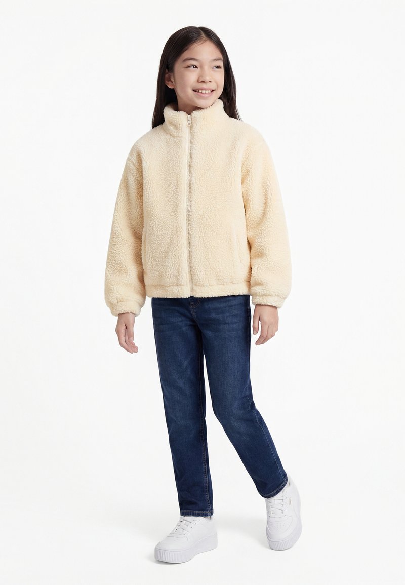 Lächendes Kind in einer cremefarbenen Fleecejacke mit Reißverschluss, blauer Jeans und weißen Turnschuhen, stehend vor weißem Hintergrund.