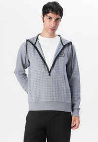 Sudadera con cremallera gris con un patrón de cuadrícula, bolsillo frontal y acentos verdes en contraste en la capucha. Tejido suave y puños elásticos.