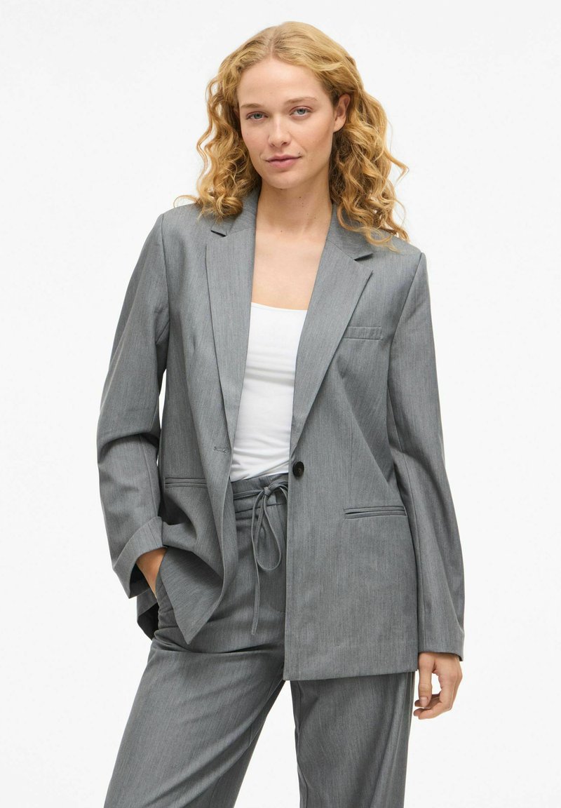 Šedý upravený blazer s jedním knoflíkem, klopovými límci a bočními kapsami, spárovaný s odpovídajícími kalhotami a bílým topem. Hladká textura látky.