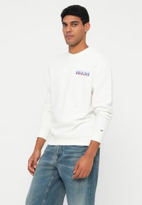 Muž oblečený v bílém svetru s dlouhým rukávem s logem "1985" a modrých džínách, stojící na jednobarevném bílém pozadí.