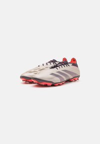 Chaussures de football gris clair avec des accents violets foncés et rouges, tige texturée, laçage central et semelle adhérente avec des crampons rouges.