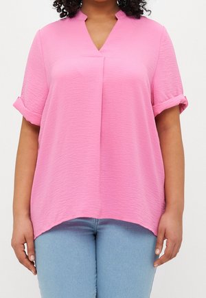 Kobieta ubrana w luźną różową bluzkę z krótkim rękawem, dekoltem w serek i podwiniętymi mankietami, w połączeniu z jasnoniebieskimi dżinsami.