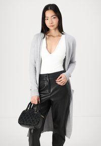 Hellgrauer Cardigan mit gerippten Bündchen, kombiniert mit schwarzen Lederhosen mit hohem Bund. In einer Hand wird eine strukturierte schwarze Handtasche gehalten.