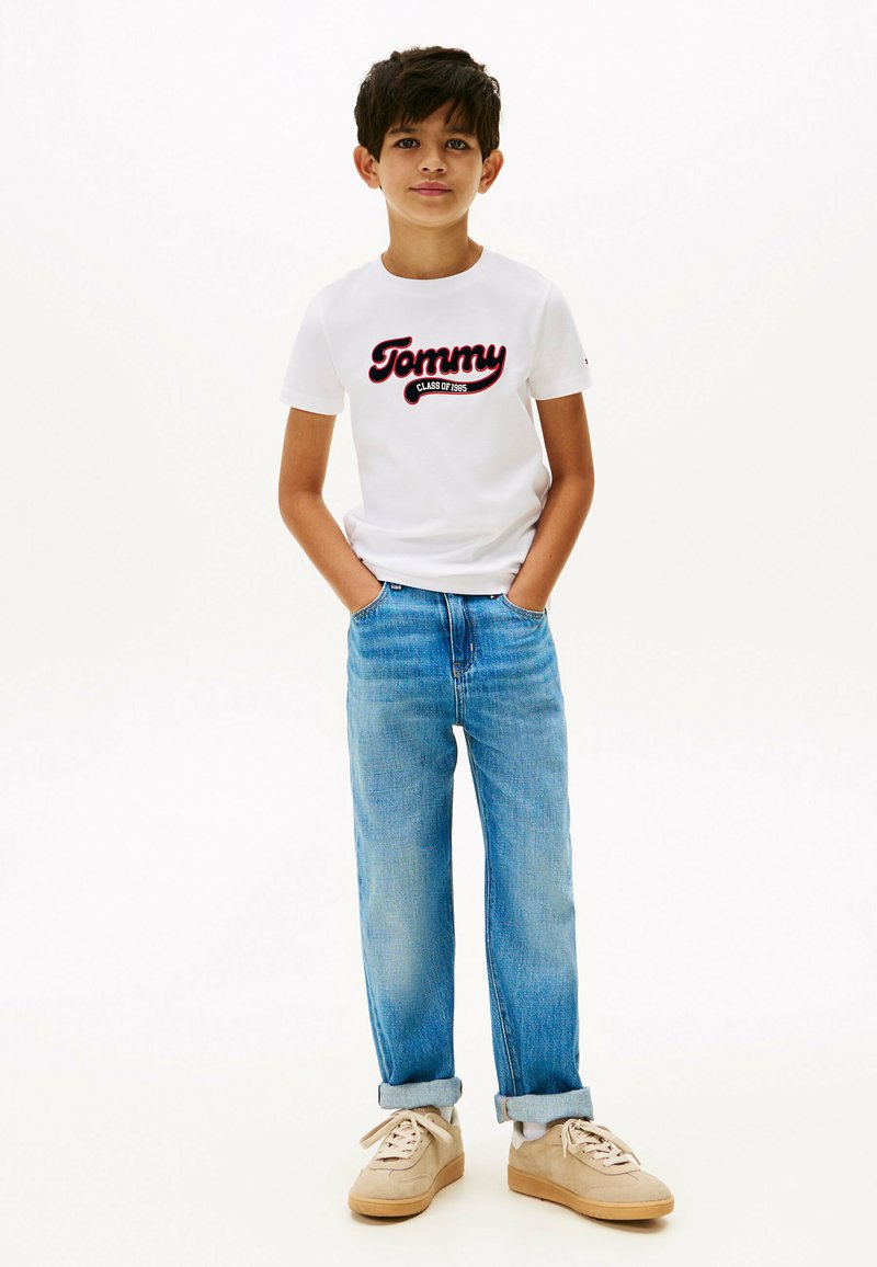 Maglietta bianca in cotone con grafica "Tommy CLASS OF 1985", jeans in denim blu e sneakers beige con dettagli stringati, in postura eretta.