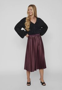 Cardigã preto curto com decote em V e fecho de botões, combinado com saia plissada vinho até ao joelho e sapatos pretos abertos à frente.