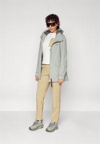 Veste de pluie gris clair avec capuche, assortie d'une chemise blanche et d'un pantalon beige. Les détails notables incluent des zips et des poches, ainsi que des chaussures texturées.