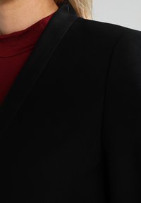 Blazer negro con una textura suave y solapas de satén, superpuesto a una blusa de color rojo oscuro. Vista en primer plano centrada en los detalles del cuello y los hombros.