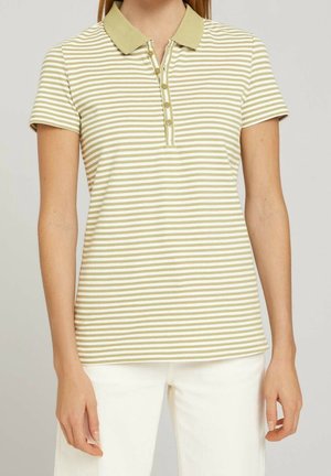 Kobieta w beżowo-białej koszulce polo w paski z krótkim rękawem, zielonym kołnierzem i guzikami, zestawiona z białymi spodniami.