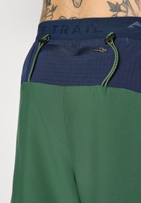Pantalones cortos verdes y azul marino con una cinturilla texturizada, que cuentan con un cierre de cordón y un pequeño bolsillo con cremallera en la parte trasera.