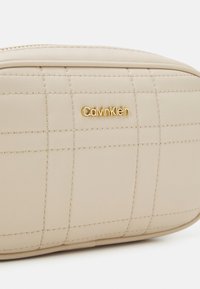 Béžová prošívaná kosmetická taštička se zlatým logem "Calvin Klein". Hladký syntetický materiál s jemnými šicími detaily a zaobleným tvarem.