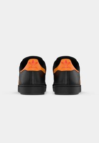 Tênis atléticos pretos com acentos laranja, apresentando uma textura de couro liso, design de perfil baixo e logotipo da Adidas na traseira do calcanhar.