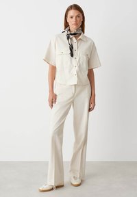 Camicia aderente a maniche corte color crema con due tasche sul petto, abbinata a pantaloni larghi dello stesso colore e scarpe da ginnastica bianche. Foulard nero a fantasia in aggiunta.