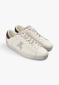 Zapatillas de piel blancas con acentos de ante beige, cordones planos y una suela de goma texturada. Presentan un logo de calavera y huesos cruzados en el lateral.