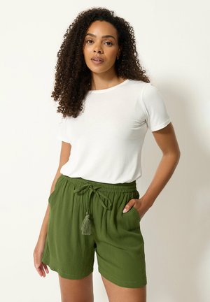 Femme aux cheveux bouclés portant une chemise blanche à manches courtes et un short vert à cordon, debout avec une main dans la poche.