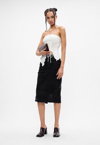 Femeie stând în picioare, îmbrăcată cu un top alb fără bretele, fustă midi texturată neagră, ținând o poșetă burgundă și pantofi cu toc închis.