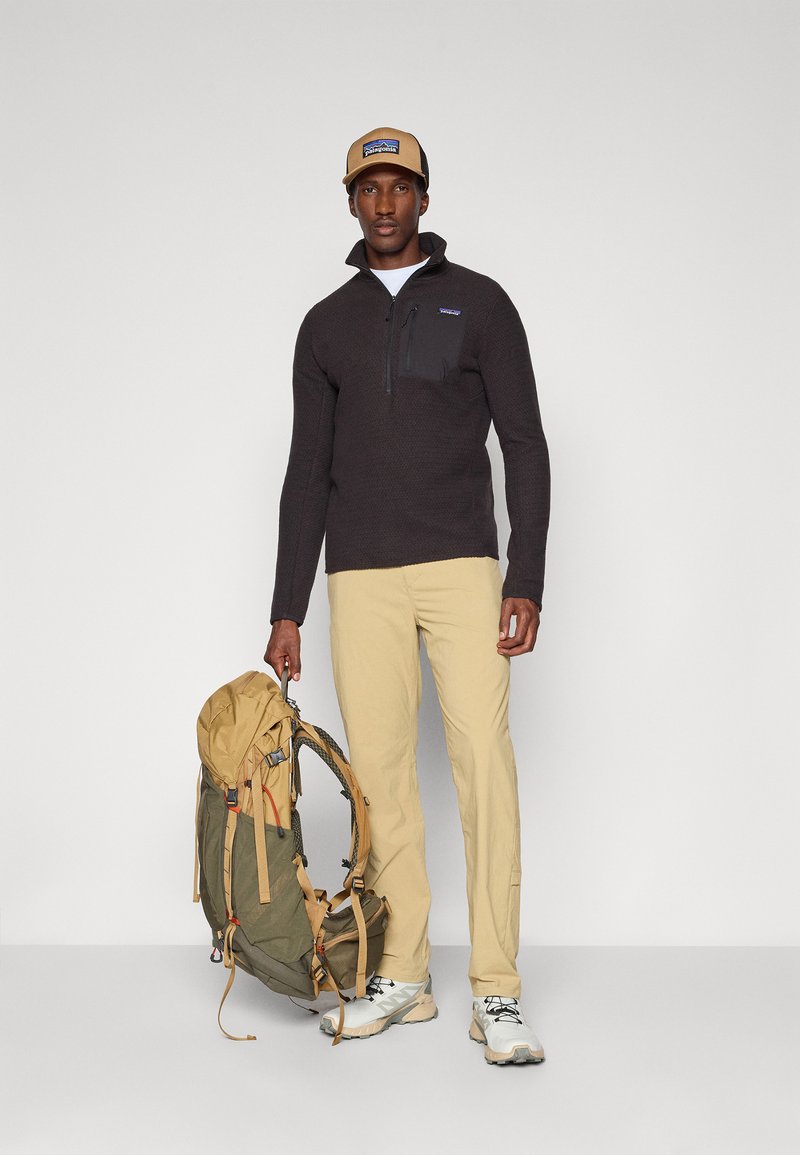 Dunkelgrauer Pullover mit Reißverschlusskragen und Brusttasche, beige Hose, beiger Hut mit Logo und grüner Rucksack mit beigen Akzenten.