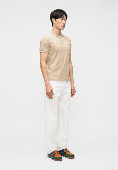 Νεαρός άνδρας με μπεζ πόλο πουκάμισο, λευκό παντελόνι και πολύχρωμα παπούτσια τύπου μοκασίνι, στέκεται μ προστά από ένα μονόχρωμο φόντο.