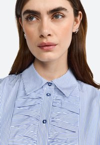 Blusa a righe blu e bianche con colletto a punta, dotata di due bottoni blu e dettagli arruffati verticali lungo il pannello frontale.