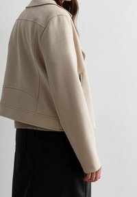 Béžová koženka s límečkem, zapínáním na zip na přední kapse a strukturovanými rameny, viděná z boku.