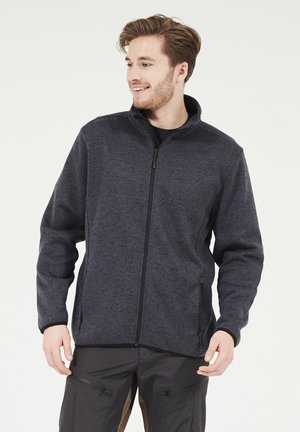 Junger Mann, der lächelt und nach rechts schaut, trägt eine dunkle Fleecejacke mit Reißverschluss und dunkle Hosen mit Reißverschlusstaschen vor einem weißen Hintergrund.