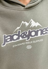 Zelena olivna kapuca s prednjo grafiko, ki prikazuje gore, blagovno znamko "jack&jones" in "STANDARD ISSUE SUPPLIES" v belem besedilu.