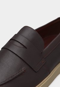 Σκούρο καφέ μοκασίνι από δέρμα με λεπτομέρεια λουριού penny και μπεζ σόλα, σε κοντινό πλάνο του πάνω μέρους και των ραφών.