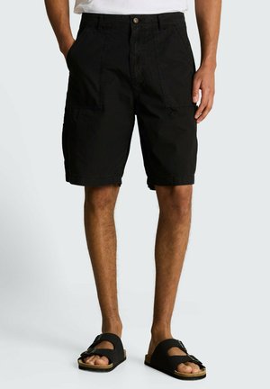 Man draagt zwarte casual shorts met voorzakken en zwarte sandalen met gesp, staat met ontspannen handen langs zijn zij.