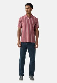 Červené tričko s krátkym rukávom a bielymi vertikálnymi pruhmi, skombinované s tmavými džínsami a šedými teniskami. Minimalistický dizajn, mäkká textúra.