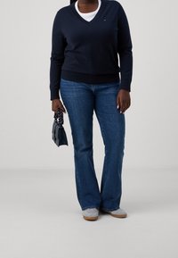 Maglione blu navy con scollo a V e maniche lunghe, abbinato a jeans blu a zampa. Tiene una piccola borsa scura. Le scarpe sono grigie con suola in gomma.