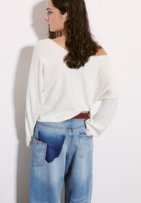 Valkoinen olkaimeton villapaita löysillä hihoilla yhdistettynä vaaleansinisiksi farkkuihin, joissa on haalistunut tumma läiskä takataskussa, sekä punainen vyö.