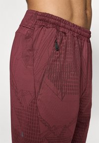 Pantalones atléticos burdeos con cintura elástica, que cuentan con un bolsillo con cremallera y una textura de patrón geométrico. Fabricados con un tejido ligero.