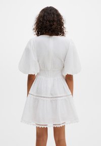 Moteris su garbanotais plaukais, vilkinti trumpą baltą suknelę su pūstomis rankovėmis ir nėrinių apdaila, stovinti atsigręžusi nugarą prieš paprastą foną.