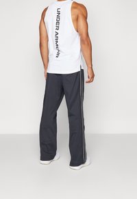 Vit ärmlös linne med "UNDER ARMOUR" tryckt vertikalt i svart; kombinerat med mörkgrå träningsbyxor med en vit sidrand.