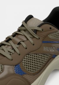 Sapatilhas castanhas e bege com painéis de malha, atacadores verdes, realces azuis e a marca "HUGO" de um lado.