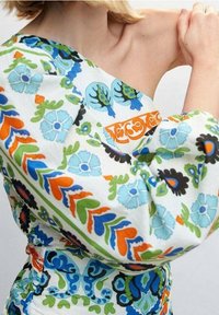 Haut asymétrique avec un motif coloré de fleurs et de feuilles en bleu, vert, orange et noir sur fond blanc. Détail de taille élastique.