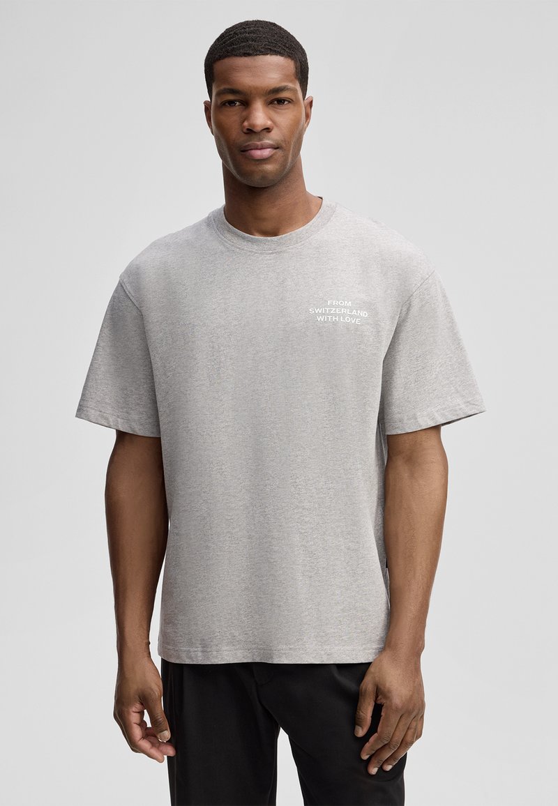 Jonge man in een grijs T-shirt met de tekst "FROM SWITZERLAND WITH LOVE", staand voor een effen lichtgrijze achtergrond.