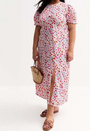 Kobieta w białej sukience midi w kwiaty z bufkowymi rękawami i rozcięciem z przodu, trzymająca plecioną torebkę, nosząca brązowe sandały wsuwane.