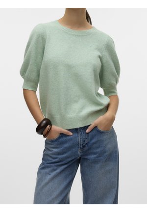 Woman wearing a light green short puff sleeve sweater with blue jeans and dark wooden bangle bracelets, hands in pockets.