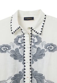 White blouse with navy floral embroidery, featuring a pointed collar and button closure. Includes decorative stitching along the collar edge.