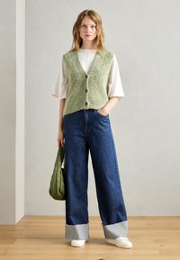 Groene, pluizige vest met een v-hals en knopen, gedragen over een wit shirt. Gecombineerd met brede, hooggepaste blauwe jeans en witte sneakers.